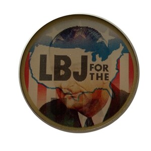 May include: A round button with a gold-colored rim. The button features a portrait of a man with the text "LBJ FOR THE" over an outline of the United States, with red, white, and blue colors.
