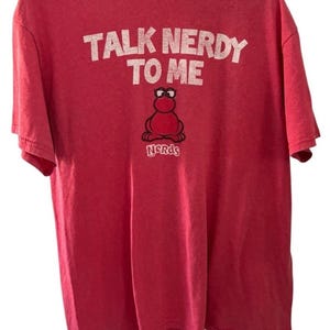 Może przedstawiać: Czerwony t-shirt z krótkim rękawem z białym napisem TALK NERDY TO ME nad grafiką przedstawiającą czerwoną postać w czarnych okularach. Pod postacią znajduje się biały napis NERDS. Koszulka posiada okrągły dekolt i standardowy krój.