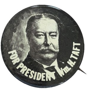 May include: A vintage campaign button featuring a black and white portrait of William H. Taft. The button reads "FOR PRESIDENT Wm. H. TAFT" in white text around the edge. The button has a pin on the back.