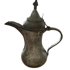 May include: Antique silver-toned metal coffee pot with a textured surface. The pot features a curved spout, a handle, and a decorative lid with a pointed finial. The design suggests a vintage or Middle Eastern style.