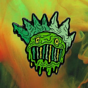 Pode incluir: Pin de esmalte verde e amarelo com um monstro de desenho animado com uma coroa de espinhos e um sorriso babante e dentado.