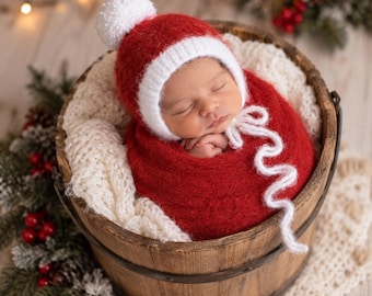 May include: A newborn infant wrapped in a deep red knitted fabric, wearing a matching red and white knitted hat with a white pom-pom and white ties. The infant is nestled inside a rustic wooden bucket lined with a soft, cream-colored knit blanket.