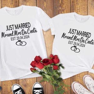 May include: Two white t-shirts with black text that says "Just Married Mr. and Mrs. Da Costa EST 29.04.2024" with a ring graphic. The shirts are laid out on a wooden surface with a bouquet of red roses in the middle.