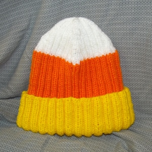 May include: A knitted hat in the shape of a candy corn. The hat features a white top, an orange middle section, and a yellow brim. The hat is made of a ribbed knit fabric.
