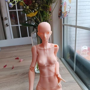 May include: A nude-colored articulated doll with long, straight, dark hair. The doll has realistic features and is posed sitting with its legs crossed. The doll is wearing a nude-colored bikini bottom.