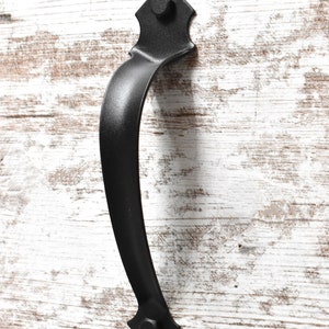 May include: Black metal door handle with a pointed top and bottom. The handle is mounted on a white wooden surface.