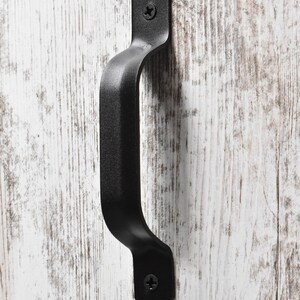 May include: Black metal door handle with a rounded rectangular shape. The handle is attached to a white wooden surface.