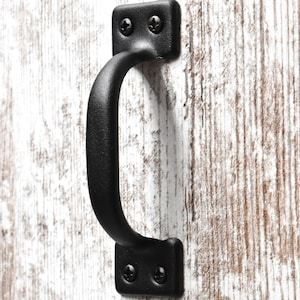 May include: Black metal door handle with a rounded rectangular shape. The handle is attached to a white wooden door.