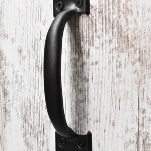 May include: Black metal cabinet pull handle with a curved design, mounted on a distressed white wooden surface. The handle is attached with screws, offering a rustic aesthetic.