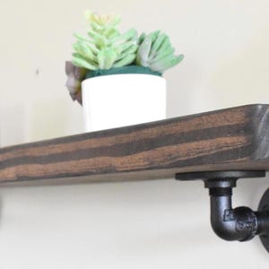 May include: A rustic, stained wood floating shelf with black pipe brackets. The shelf holds a small white pot with a green plant and a blue and white candle holder. A decorative and functional home decor item.
