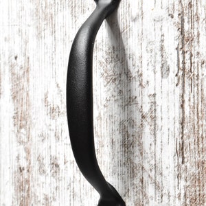 May include: A black, curved cabinet pull with a textured finish. The handle is mounted with screws at each end, set against a white-washed wooden surface. This hardware is suitable for cabinets, drawers, or furniture.