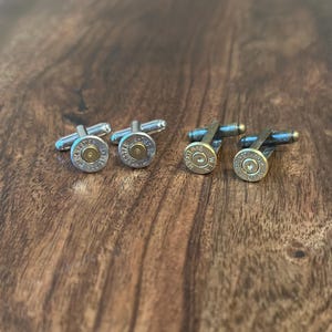 May include: Four cufflinks with a bullet casing design. Two pairs of cufflinks are visible, each with a silver or bronze finish. The bullet casings have a gold-coloured centre and are set against a wooden background.