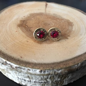 May include: A pair of gold-coloured stud earrings with a red gemstone centre. The earrings are set in a casing with the text "WIN" and "LUGER" engraved around the gemstone. The earrings are displayed on a wooden slice.