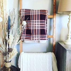 May include: A wooden ladder displays two blankets. The top blanket has a plaid pattern in burgundy, pink, and white. The bottom blanket is white with a cable knit design. A vase with decorative branches and a lamp are also visible.