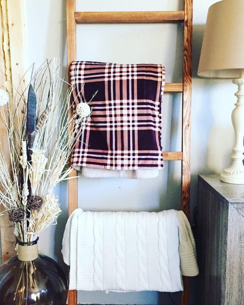 May include: A wooden ladder displays two blankets. The top blanket has a plaid pattern in burgundy, pink, and white. The bottom blanket is white with a cable knit design. A vase with decorative branches and a lamp are also visible.
