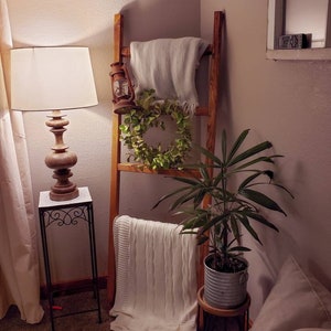 May include: A wooden ladder leans against a wall, adorned with a light-colored blanket, a green wreath, and a vintage lantern. A potted plant sits on a small wooden stool. A lamp with a wooden base and white shade sits on a black metal side table.