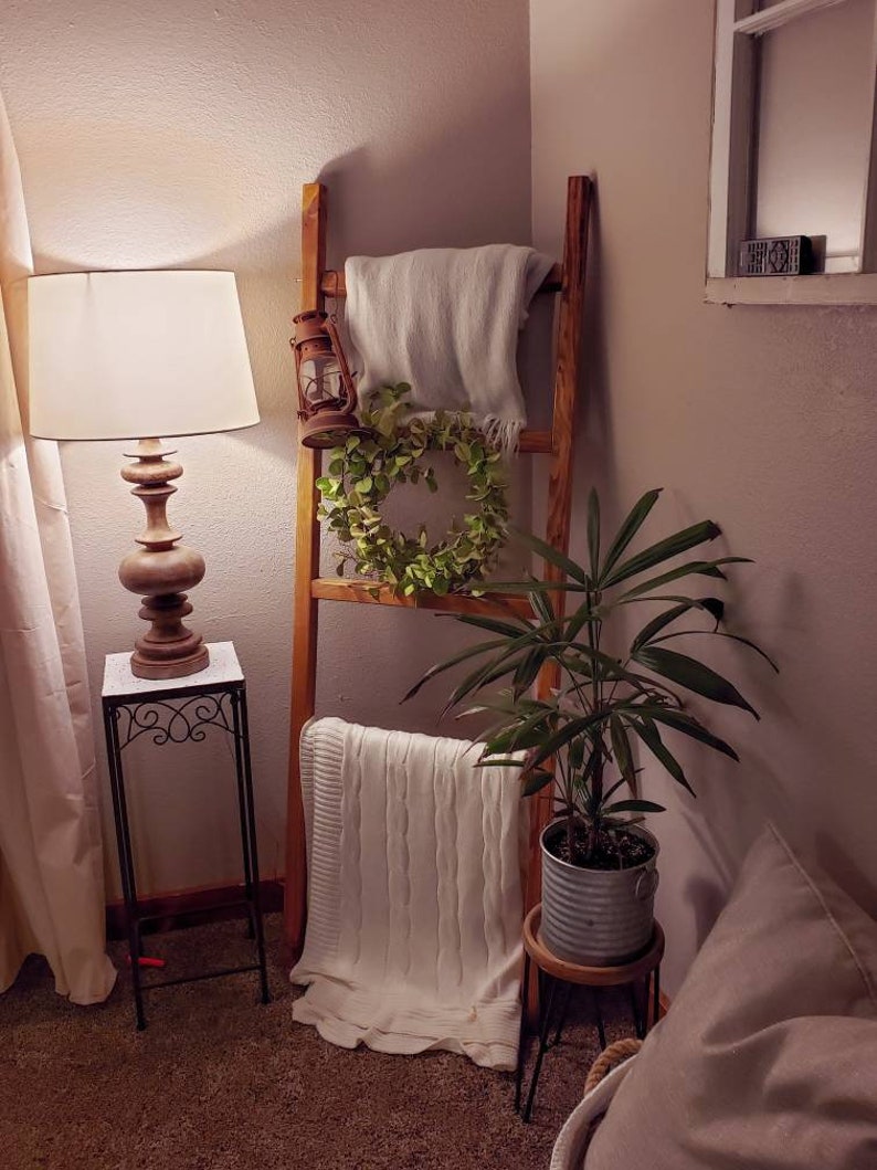 May include: A wooden ladder leans against a wall, adorned with a light-colored blanket, a green wreath, and a vintage lantern. A potted plant sits on a small wooden stool. A lamp with a wooden base and white shade sits on a black metal side table.