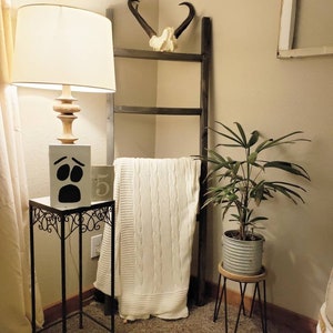 May include: A decorative corner arrangement featuring a wooden ladder, a white cable-knit blanket, and a small side table with a lamp. The lamp has a beige shade and a wooden base. A small plant in a metal pot sits on a wooden stand.