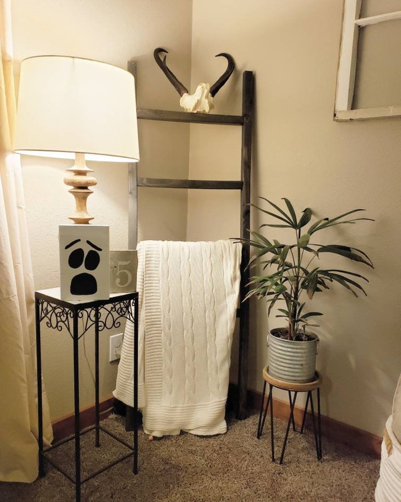 May include: A decorative corner arrangement featuring a wooden ladder, a white cable-knit blanket, and a small side table with a lamp. The lamp has a beige shade and a wooden base. A small plant in a metal pot sits on a wooden stand.
