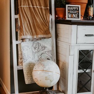 May include: A white wooden ladder displays a folded tan blanket and a patterned quilt. A vintage globe sits on a small, distressed wooden stool. A sign that says "Happy HalloWine" is on a white cabinet.
