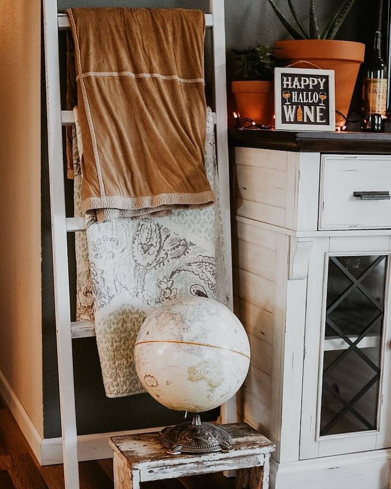 May include: A white wooden ladder displays a folded tan blanket and a patterned quilt. A vintage globe sits on a small, distressed wooden stool. A sign that says "Happy HalloWine" is on a white cabinet.