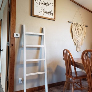 May include: A white wooden ladder leans against a beige wall. A framed sign with the text "If you want to change the world, go home and love your family" hangs above the ladder. A macrame wall hanging and wooden chairs are also visible.