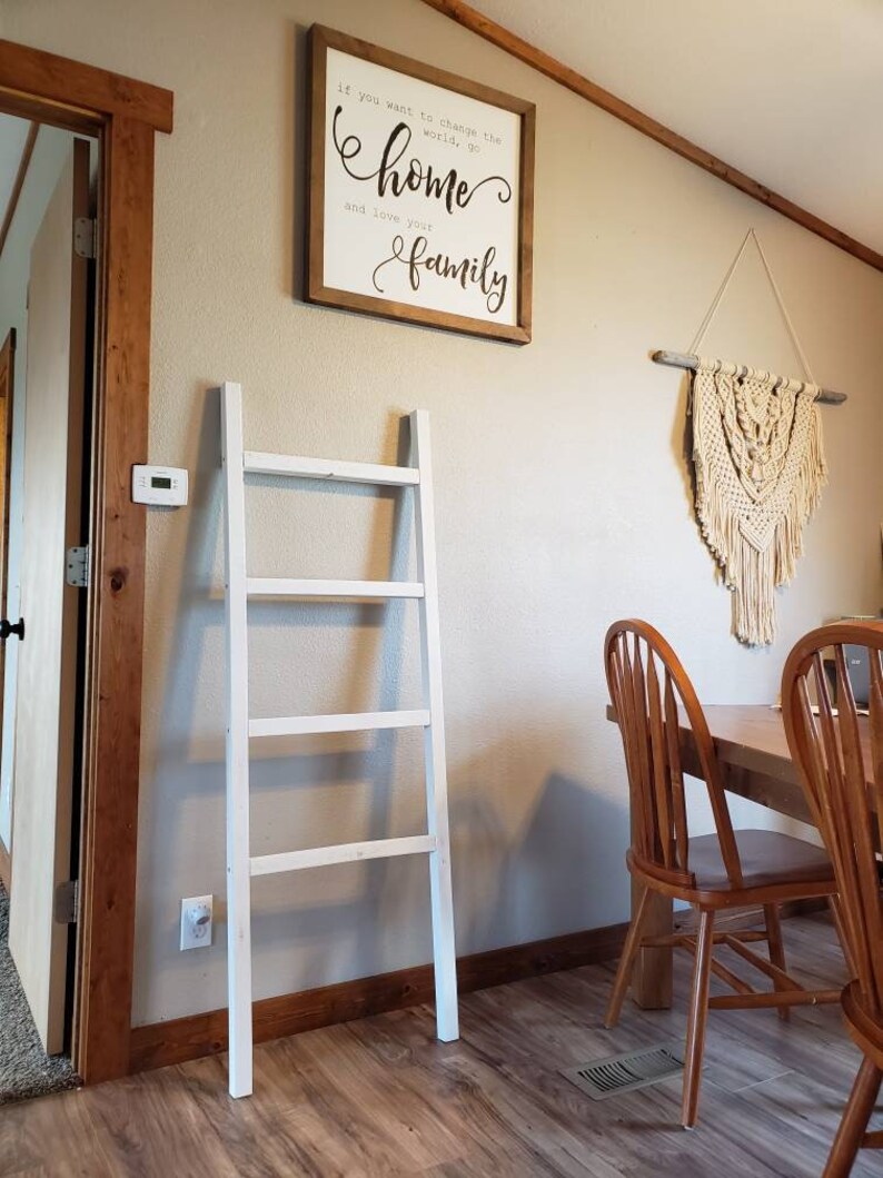 May include: A white wooden ladder leans against a beige wall. A framed sign with the text "If you want to change the world, go home and love your family" hangs above the ladder. A macrame wall hanging and wooden chairs are also visible.