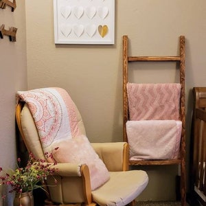 May include: A nursery scene featuring a cream-colored rocking chair with a pink pillow and blanket. A wooden ladder holds pink blankets. A framed artwork with white and gold hearts hangs on the wall. A wooden side table holds a vase of flowers.