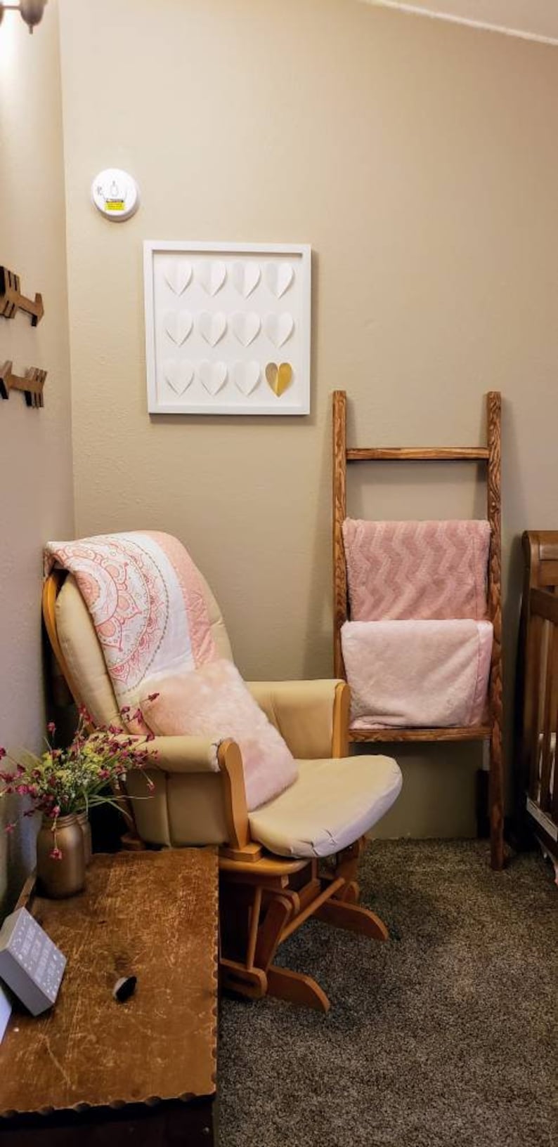May include: A nursery scene featuring a cream-colored rocking chair with a pink pillow and blanket. A wooden ladder holds pink blankets. A framed artwork with white and gold hearts hangs on the wall. A wooden side table holds a vase of flowers.