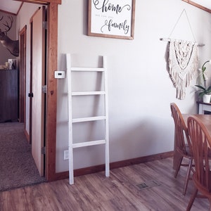 May include: A white wooden decorative ladder leans against a wall. A framed sign with the words "home and love your family" hangs above the ladder. A macrame wall hanging and wooden chairs are also visible.