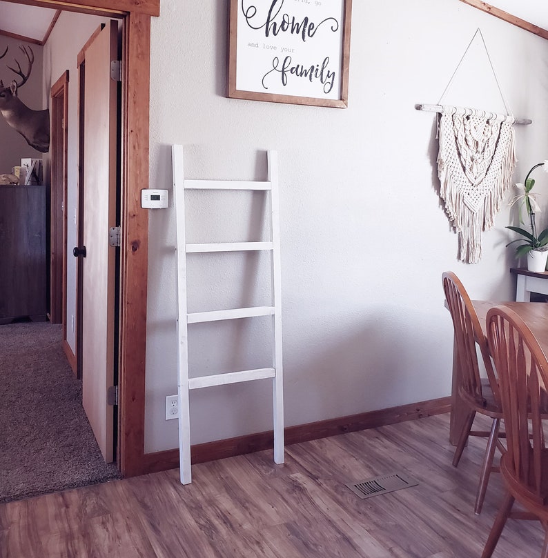 May include: A white wooden decorative ladder leans against a wall. A framed sign with the words "home and love your family" hangs above the ladder. A macrame wall hanging and wooden chairs are also visible.