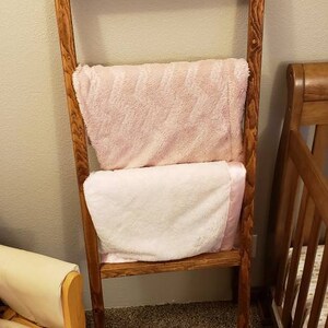 May include: A wooden blanket ladder with two blankets draped over it. The blankets are a light pink and white. The ladder is made of stained wood and is leaning against a wall. A crib and chair are in the background.