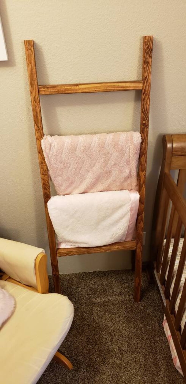 May include: A wooden blanket ladder with two blankets draped over it. The blankets are a light pink and white. The ladder is made of stained wood and is leaning against a wall. A crib and chair are in the background.