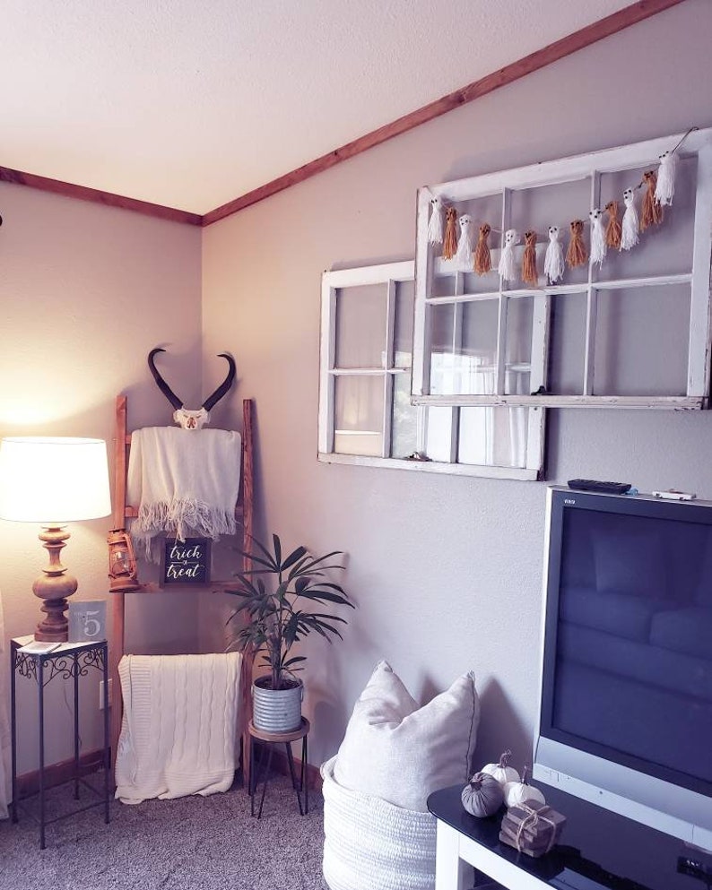 May include: A cozy interior scene with Halloween decorations. A white window frame is adorned with a ghost garland. A wooden ladder displays a skull, blanket, and a sign that reads "trick or treat". A lamp and a plant add to the decor.