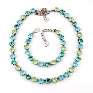 May include: A necklace and bracelet set featuring round, colorful gemstones in shades of teal, light blue, and green. The jewelry has a silver-toned chain and clasp. The necklace and bracelet are displayed on a white background.