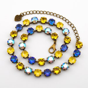 May include: A necklace featuring round, faceted crystals in alternating shades of vibrant yellow, deep blue, and iridescent white, set in a bronze-toned metal chain. The piece includes a lobster claw clasp and a small heart-shaped charm engraved with the text Made With Love.