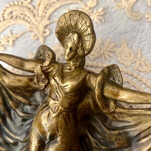 May include: A bronze-colored sculpture of a woman in a flowing gown and hat, arms outstretched. The figure has detailed facial features and is posed in a graceful, dancing stance. The sculpture is likely a decorative item.