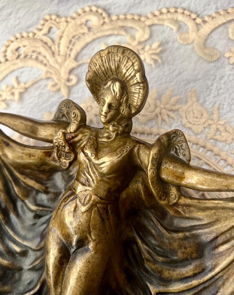 May include: A bronze-colored sculpture of a woman in a flowing gown and hat, arms outstretched. The figure has detailed facial features and is posed in a graceful, dancing stance. The sculpture is likely a decorative item.