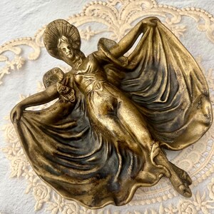 May include: A bronze-colored decorative dish in the shape of a woman in a flowing gown. The woman is wearing a hat and has her arms outstretched, holding the fabric of her dress. The dish has intricate details and a vintage aesthetic.