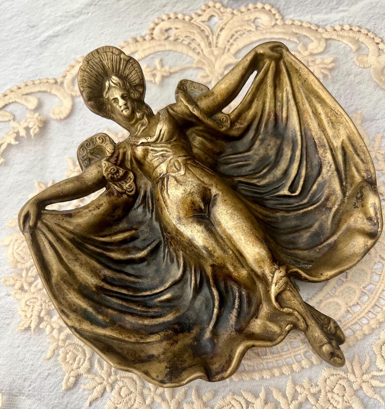 May include: A bronze-colored decorative dish in the shape of a woman in a flowing gown. The woman is wearing a hat and has her arms outstretched, holding the fabric of her dress. The dish has intricate details and a vintage aesthetic.