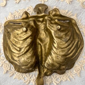 May include: A vintage brass decorative dish with a unique, organic shape. The dish has a central stem and two curved compartments, with a textured surface and small, raised details. The brass has a warm, aged patina.