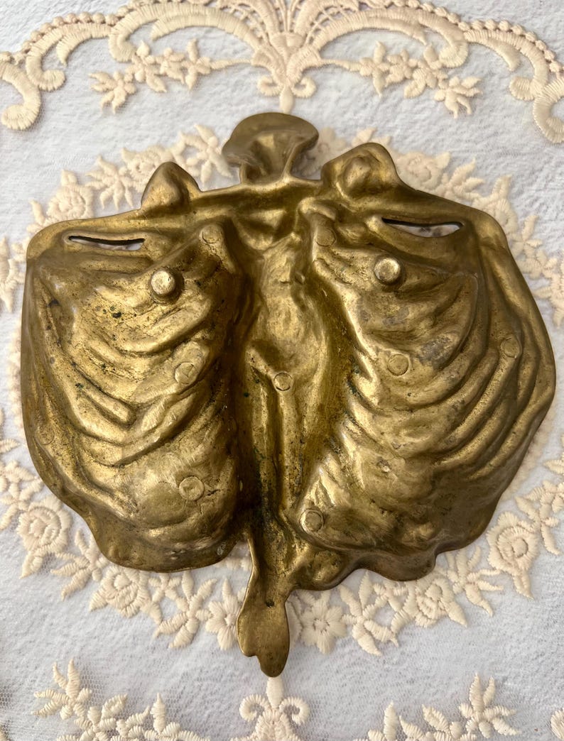 May include: A vintage brass decorative dish with a unique, organic shape. The dish has a central stem and two curved compartments, with a textured surface and small, raised details. The brass has a warm, aged patina.