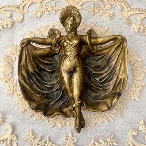 May include: A bronze-colored wall sculpture of a woman with outstretched arms, wearing a hat and flowing gown. The figure is detailed with intricate folds in the fabric and is mounted on a white surface.