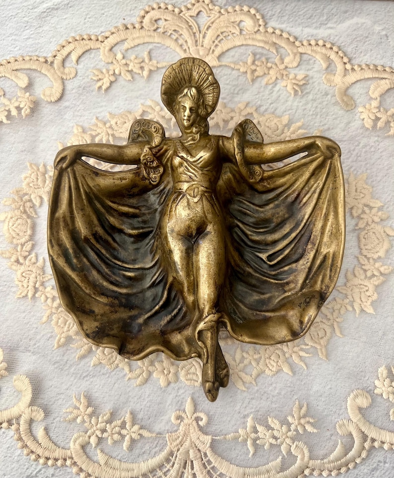 May include: A bronze-colored wall sculpture of a woman with outstretched arms, wearing a hat and flowing gown. The figure is detailed with intricate folds in the fabric and is mounted on a white surface.
