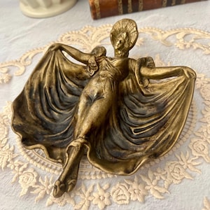 May include: A bronze-toned Art Deco figurine of a woman with outstretched arms, her flowing dress forming a shallow dish. The figure has a detailed headdress and is set against a lace tablecloth.