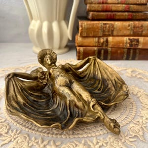 May include: A bronze-toned decorative dish featuring a reclining female figure with flowing drapery. The dish has a shallow, curved shape and is likely made of metal. The figure is detailed with a hat and floral accents.