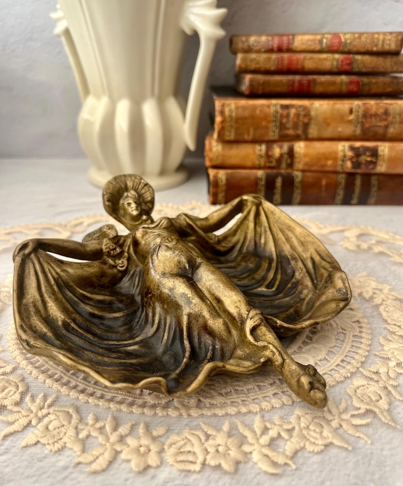 May include: A bronze-toned decorative dish featuring a reclining female figure with flowing drapery. The dish has a shallow, curved shape and is likely made of metal. The figure is detailed with a hat and floral accents.