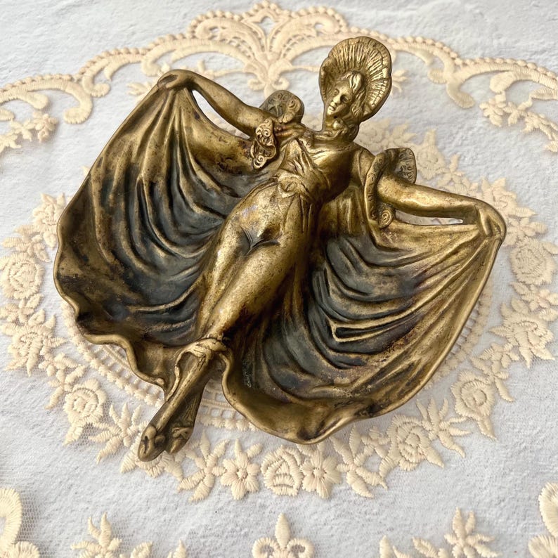 May include: A vintage brass figurine of a dancing woman with outstretched arms, holding the edges of her flowing gown. The figure has a detailed face and headdress. The brass has a tarnished, antique finish, and the figurine is likely a decorative item.