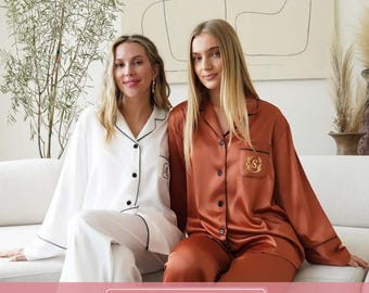 May include: Two-piece pyjama sets featuring long-sleeved button-down shirts with collars and matching trousers. One set is solid white with black piping, and the other is solid rust-orange with black piping and a gold embroidered letter S.