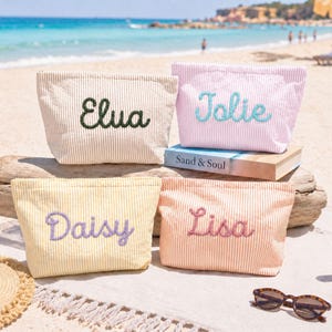 May include: Four rectangular zippered pouches made of striped fabric in beige, pink, yellow, and peach. Each pouch features a name embroidered in cursive script: Elua in dark green, Jolie in light blue, Daisy in purple, and Lisa in pink.